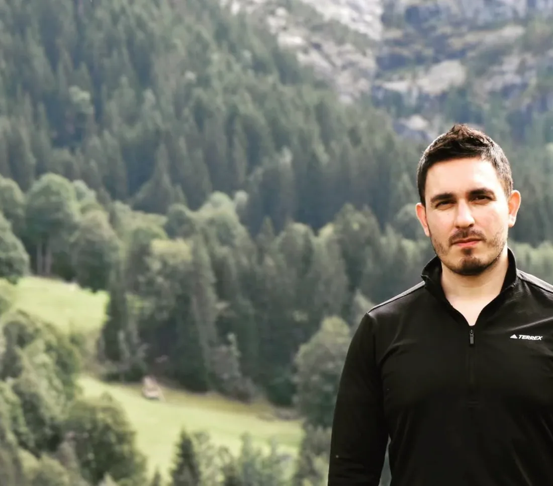 Felipe in a black zip-neck top in front of a forested mountainside in Austria