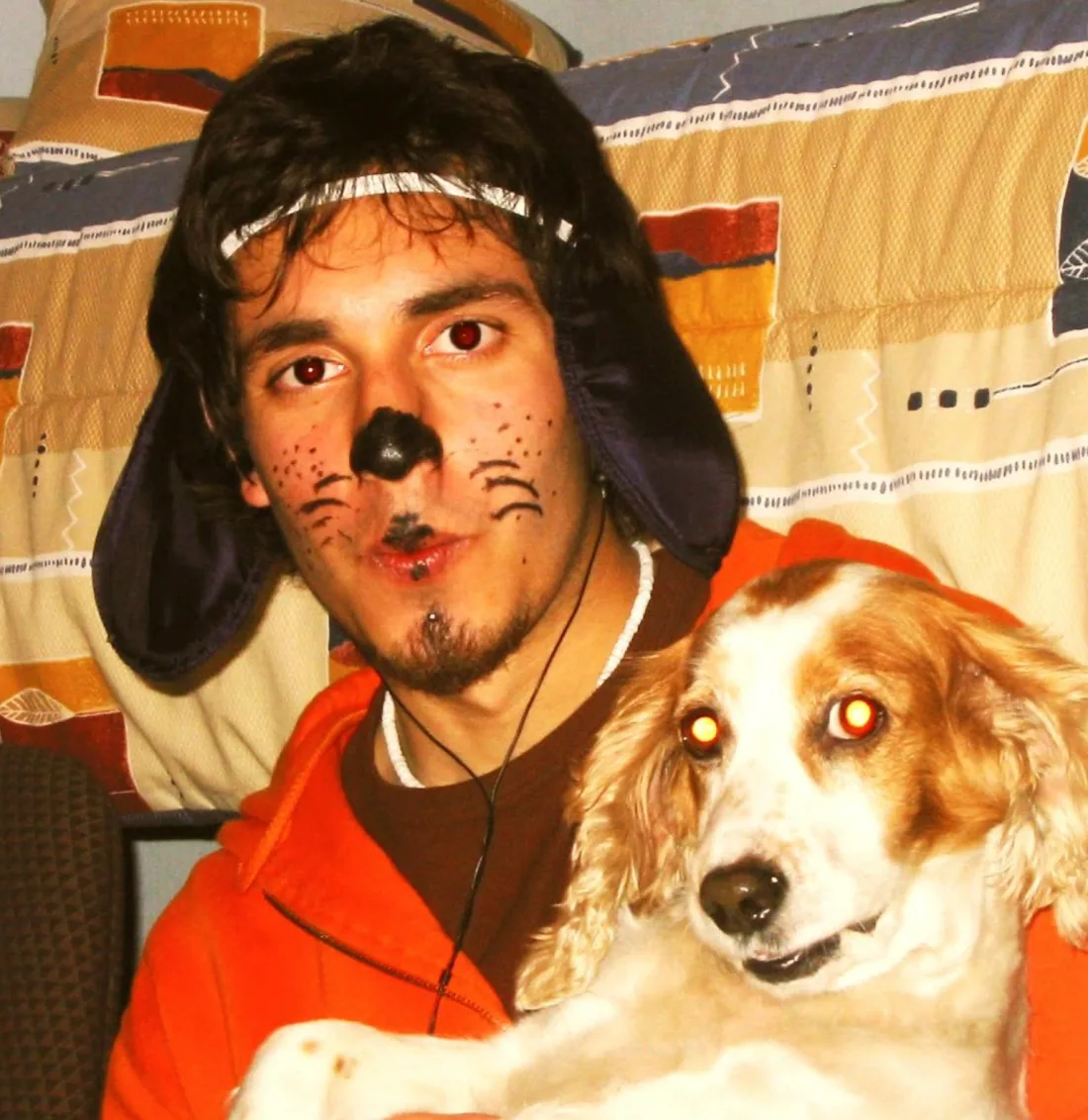 Felipe with a painted black nose and whiskers, wearing a dark hat with ear flaps and an orange jacket, holding a brown-and-white spaniel